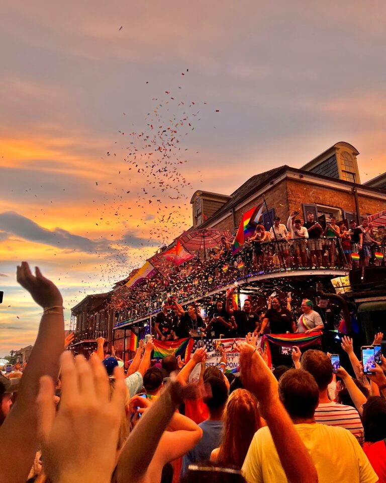 People celebrating Pride in the French Quarter from balconies and the street below as glitter floats down against the sunset