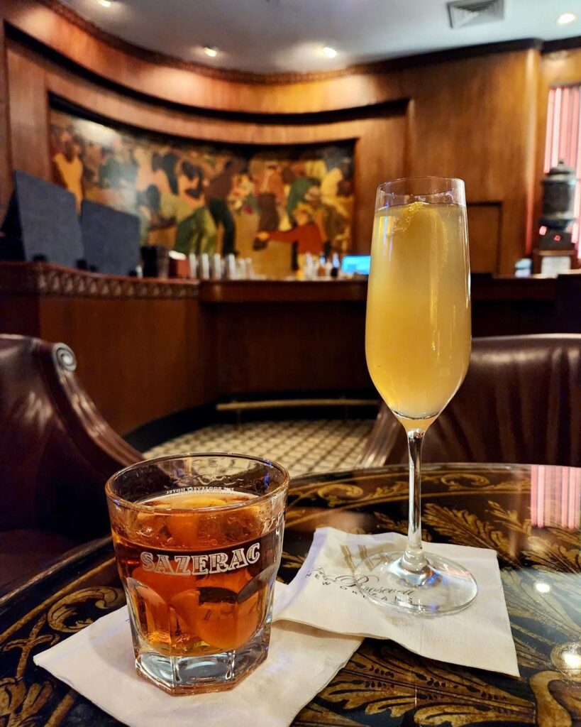 Photo of a sazerac and French 75 cocktail, each with fresh lemon garnish on top of one of The Sazerac Bar's beautiful tables, surrounded by leather club chairs, with the stately bar's surroundings in the background.