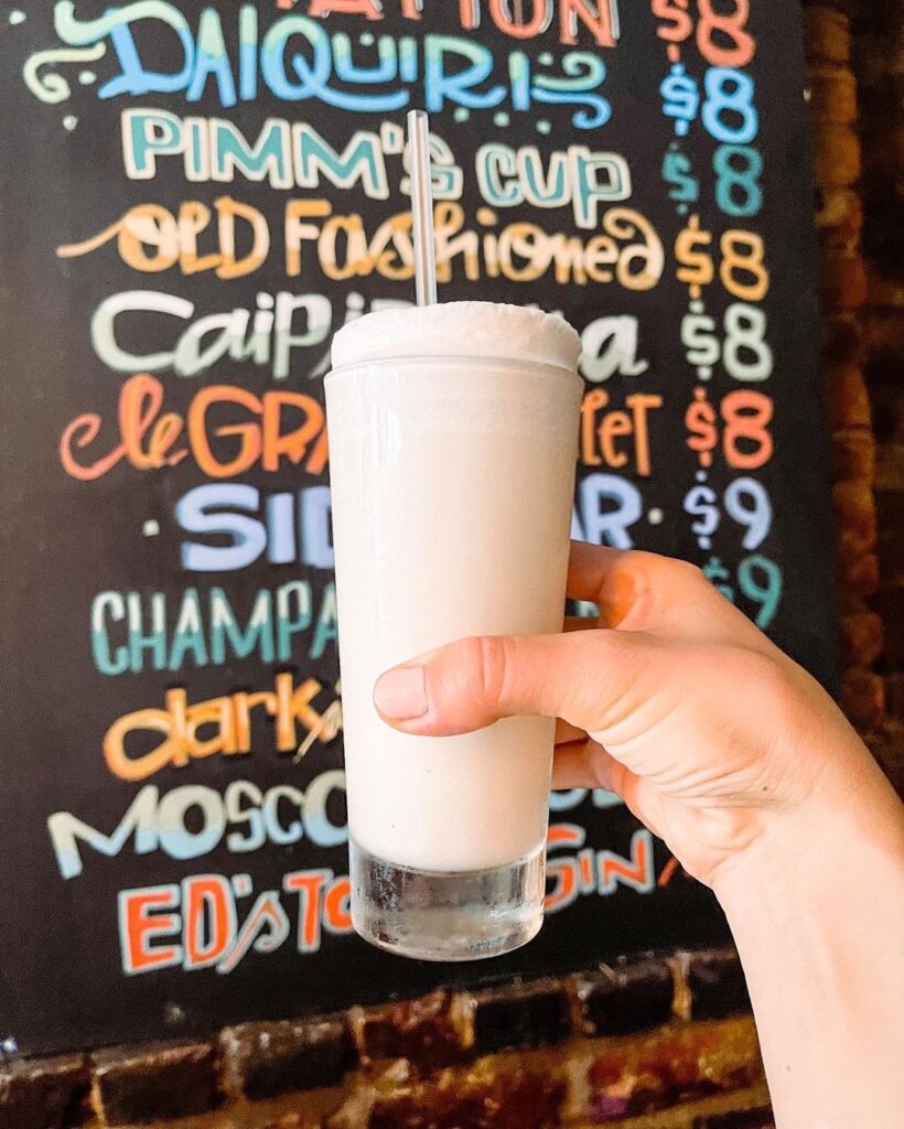 Photo of a Ramos Gin Fizz cocktail held in front of Bar Tonique's colorful hand-lettered chalkboard sign showcasing their specialty drinks